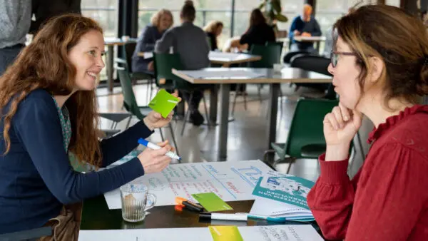 Twee personen aan een tafel in gesprek met op tafel een groot wit vel waar op geschreven wordt.