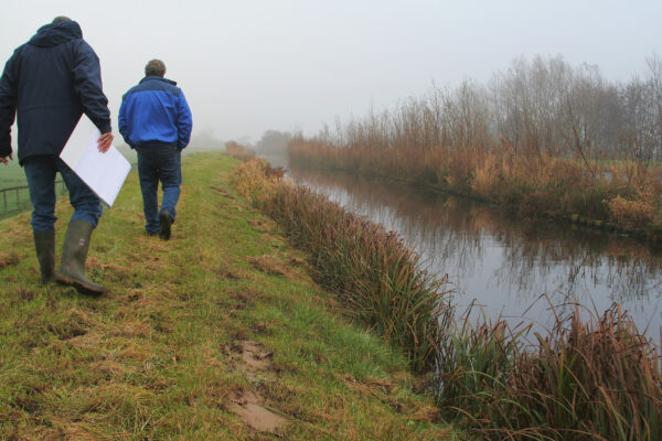 Twee personen lopen op een kleine dijk langs een watergang.